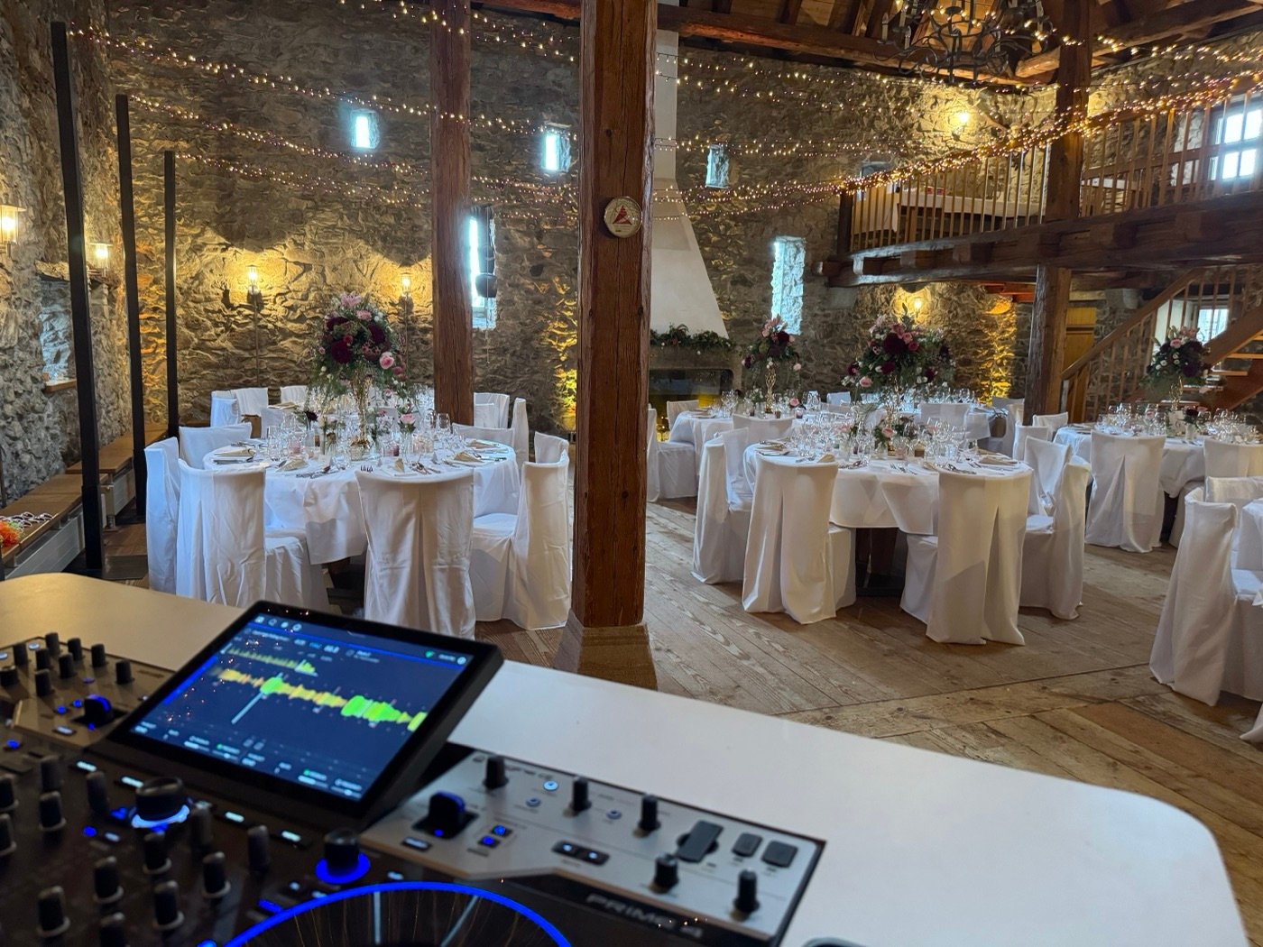 Blick vom DJ-Pult in die Scheune des Raben: Natursteinwände, Holzbalken, Galerie, Lichterketten und runde Tische mit Blumenschmuck