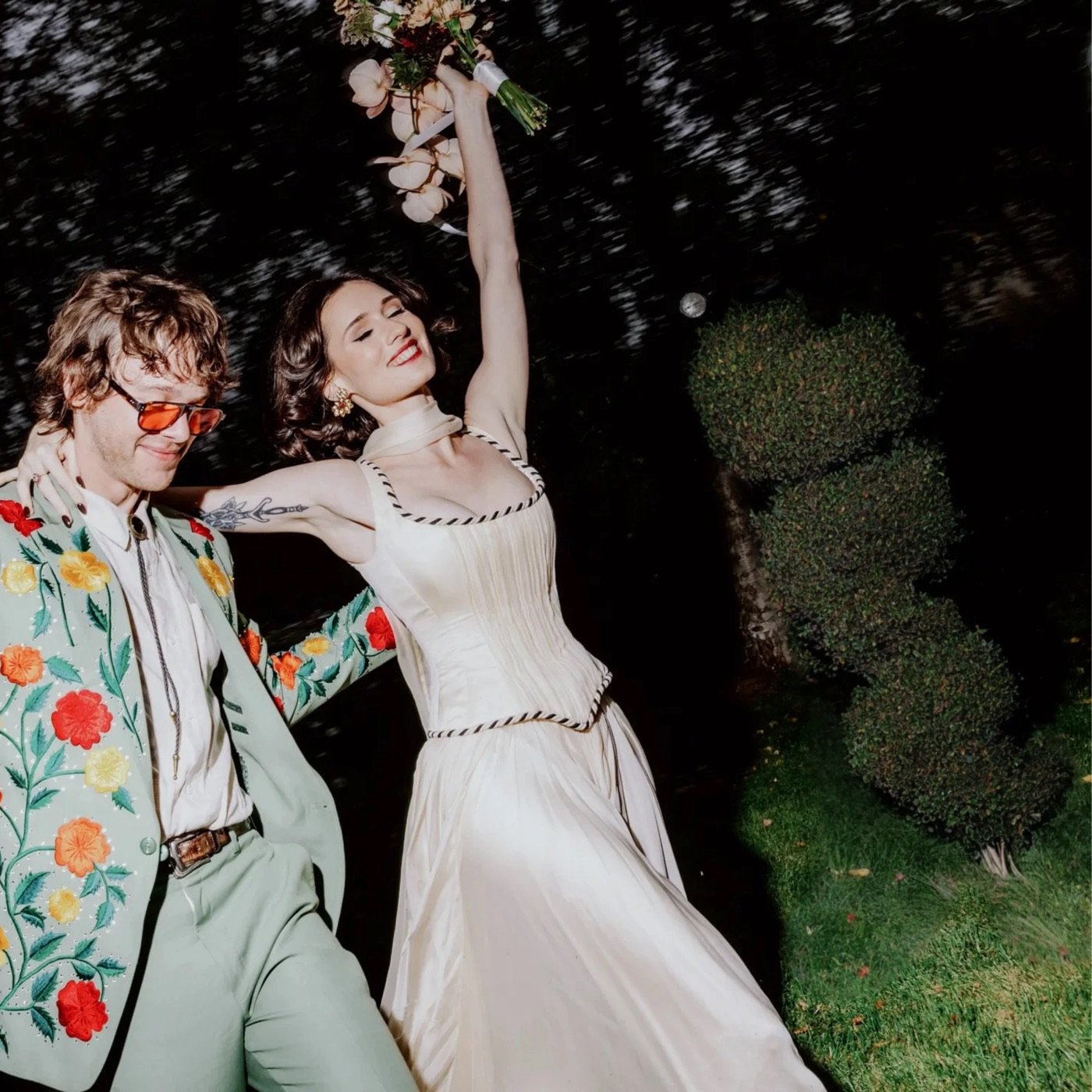 A wedding couple celebrating in a garden at night, the bride holding her bouquet high